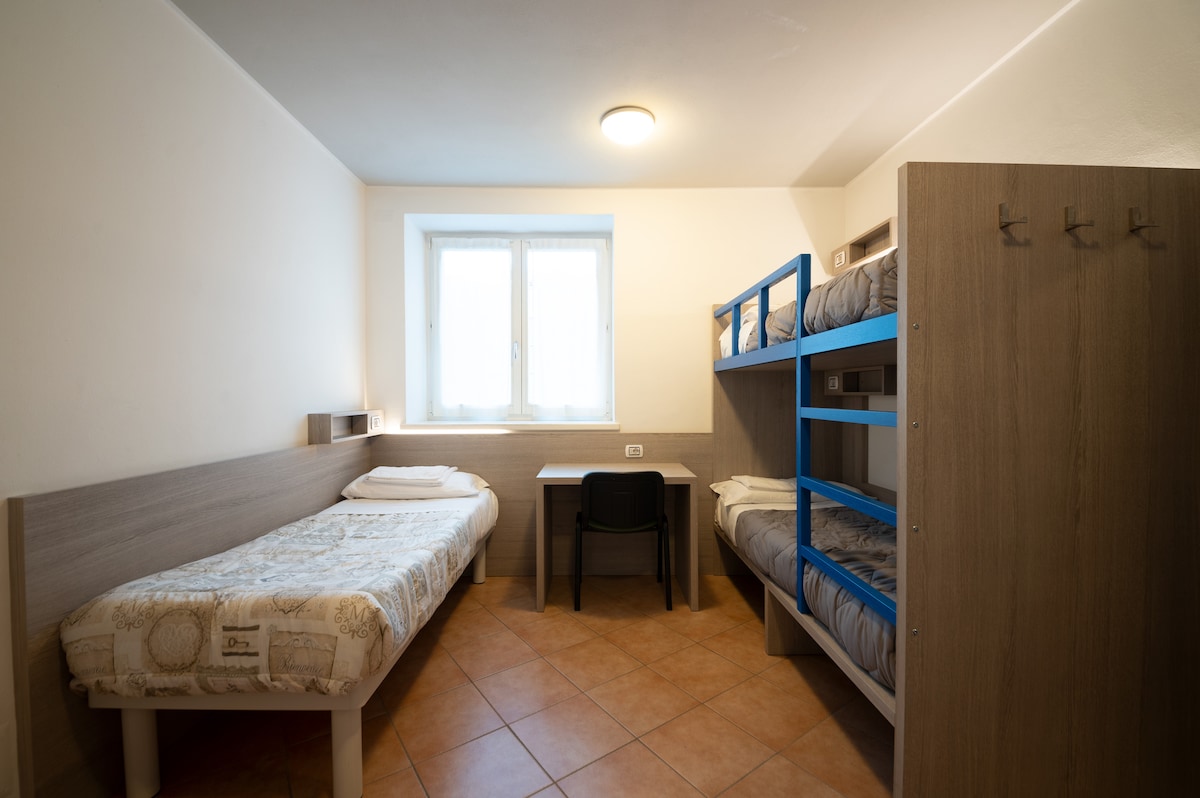 The room features a bunk bed along with a single bed, providing ample sleeping space. A simple desk and chair are positioned in the room, complementing the minimalist design. Natural light enters through the window, and warm tile flooring adds to the cozy environment.