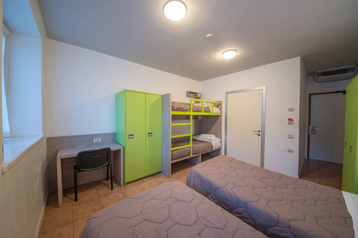 A recently renovated room is designed for families, featuring two single beds and a bunk bed. A desk is positioned against one wall, and cabinets in a bright green color add a modern touch. Natural light enters through a window, illuminating the space.