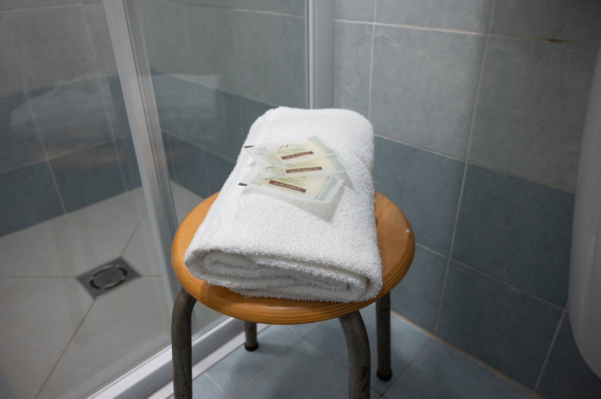 A white towel is neatly folded on a wooden stool within a tiled shower space. Small toiletries are placed beside the towel, all arranged in a clean and accessible manner.