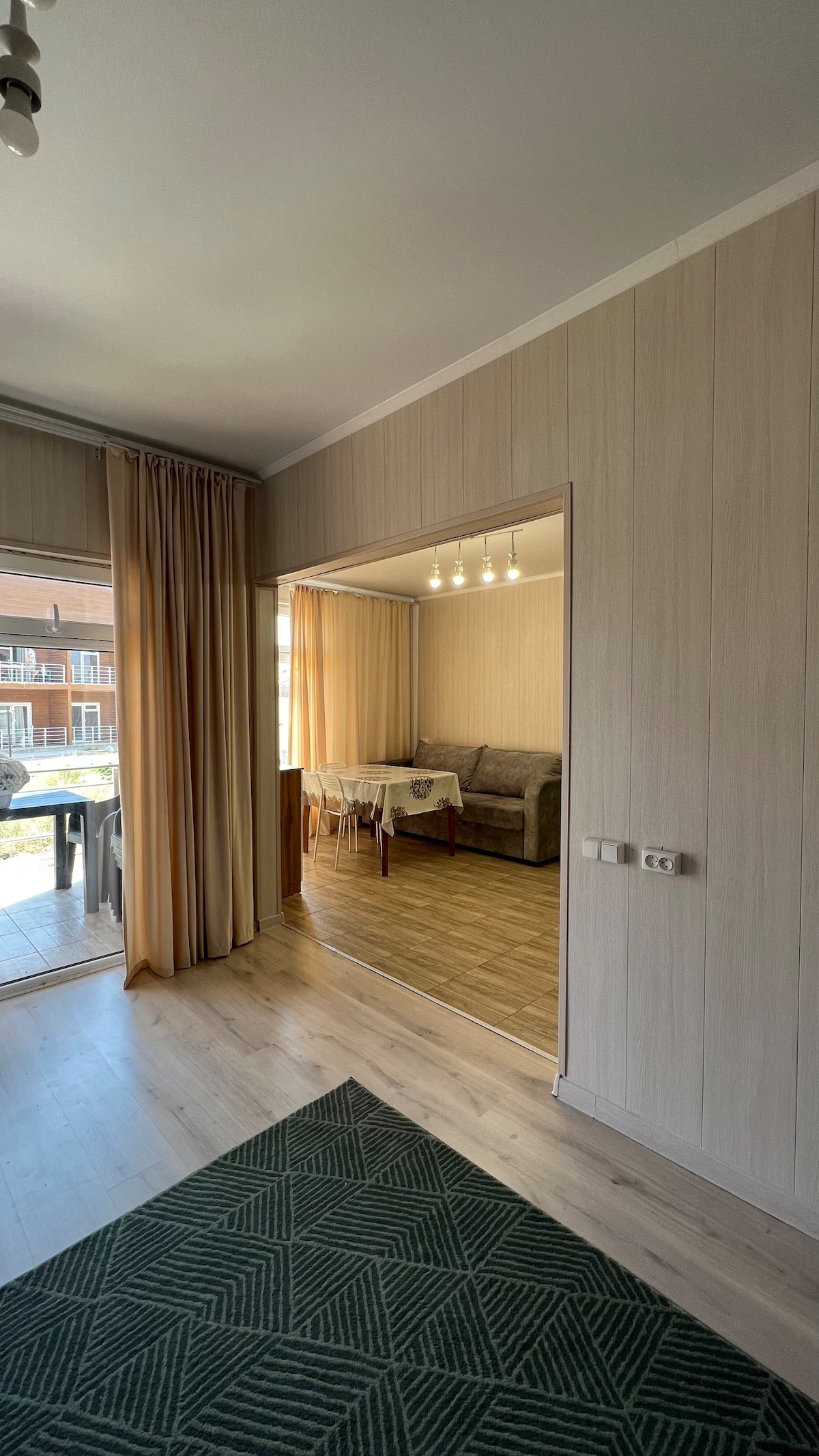 An open living space is revealed through a doorway, featuring a cozy seating area with a sofa and a dining table set for four. Natural light fills the room, enhanced by soft, neutral-coloured curtains hanging beside wide windows.