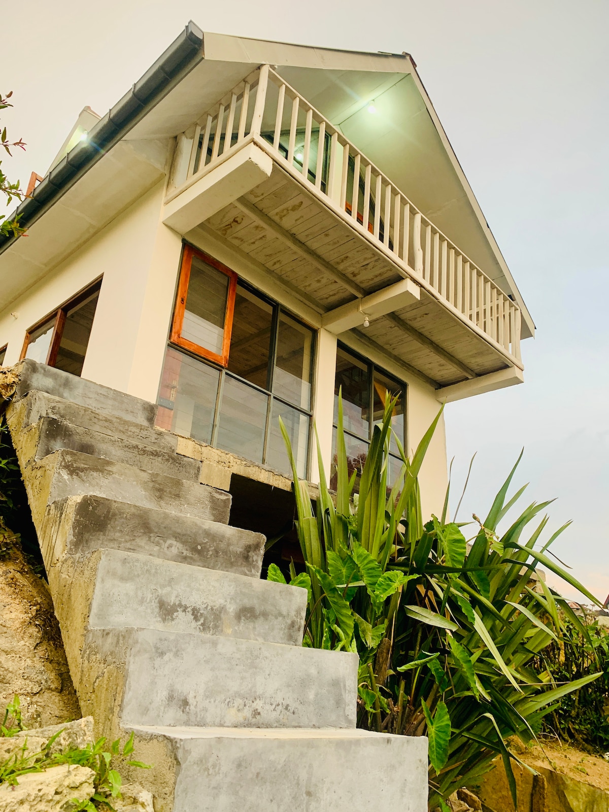 A two-story cabin is seen from a lower angle, emphasizing its sloping exterior and balcony. Stone steps lead up to the entrance, flanked by green plants. Large glass windows create an inviting connection to the outdoors.