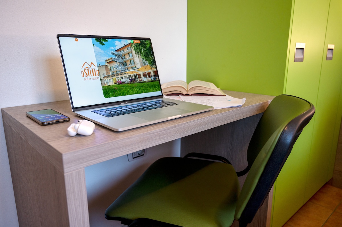 A workspace is presented featuring a wooden desk with a laptop, a closed book, and a mobile phone. Headphones are positioned beside the items. A green wall complements a nearby cabinet with secured compartments. A chair is angled towards the desk.