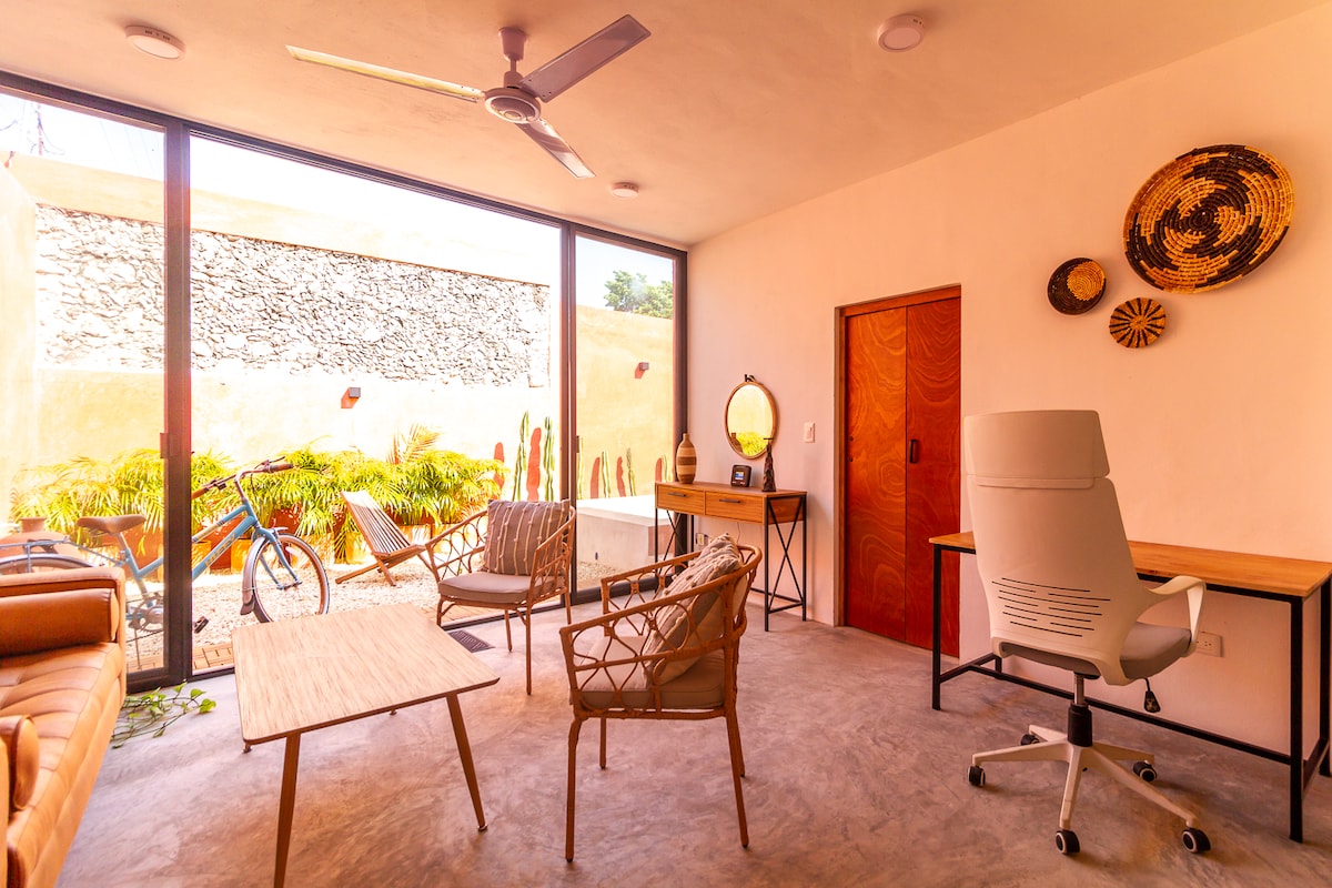 A well-lit living space features a mix of seating options, including a stylish chair and a comfortable sofa. A wooden table occupies the center, with a workspace positioned nearby. Large glass doors open to a serene outdoor area, framed by lush greenery and sculptural elements.