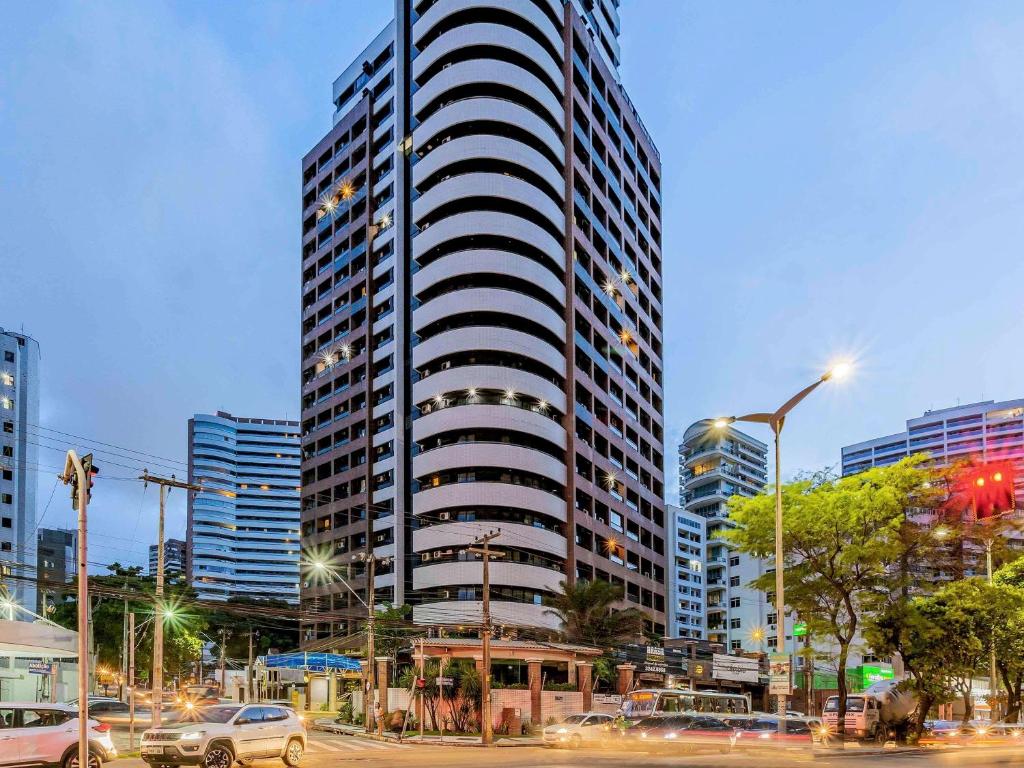 The exterior of the tall building showcases a modern architectural design with distinct circular balconies. Surrounding streets and nearby buildings are visible, featuring streetlights illuminating the area in the evening. A blend of urban elements, including vehicles and greenery, enhances the city's vibrant atmosphere.