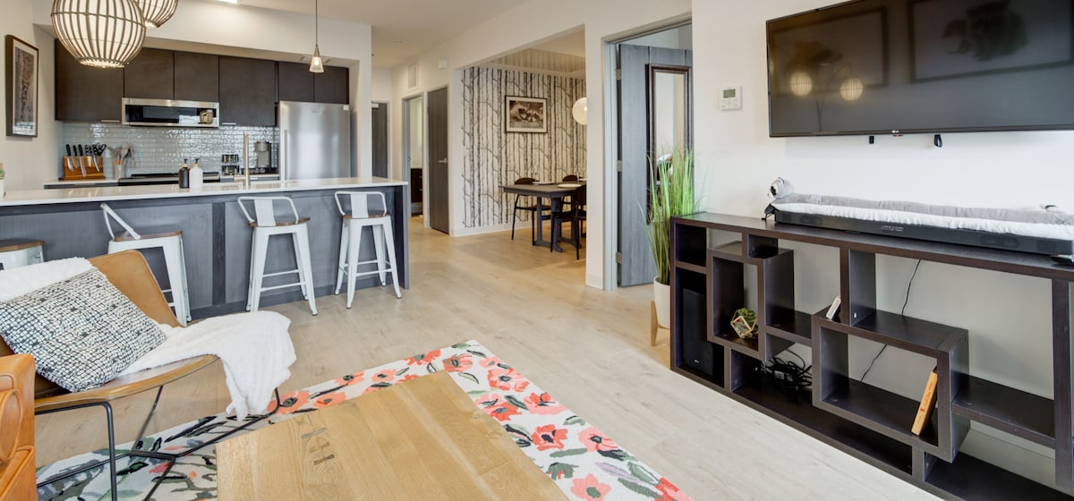 A modern living space features a sleek kitchen with dark cabinetry and stainless steel appliances. Light wood floors enhance the open layout, while contemporary furnishings include a leather chair and a decorative rug. Natural light filters in, highlighting the stylish decor throughout.