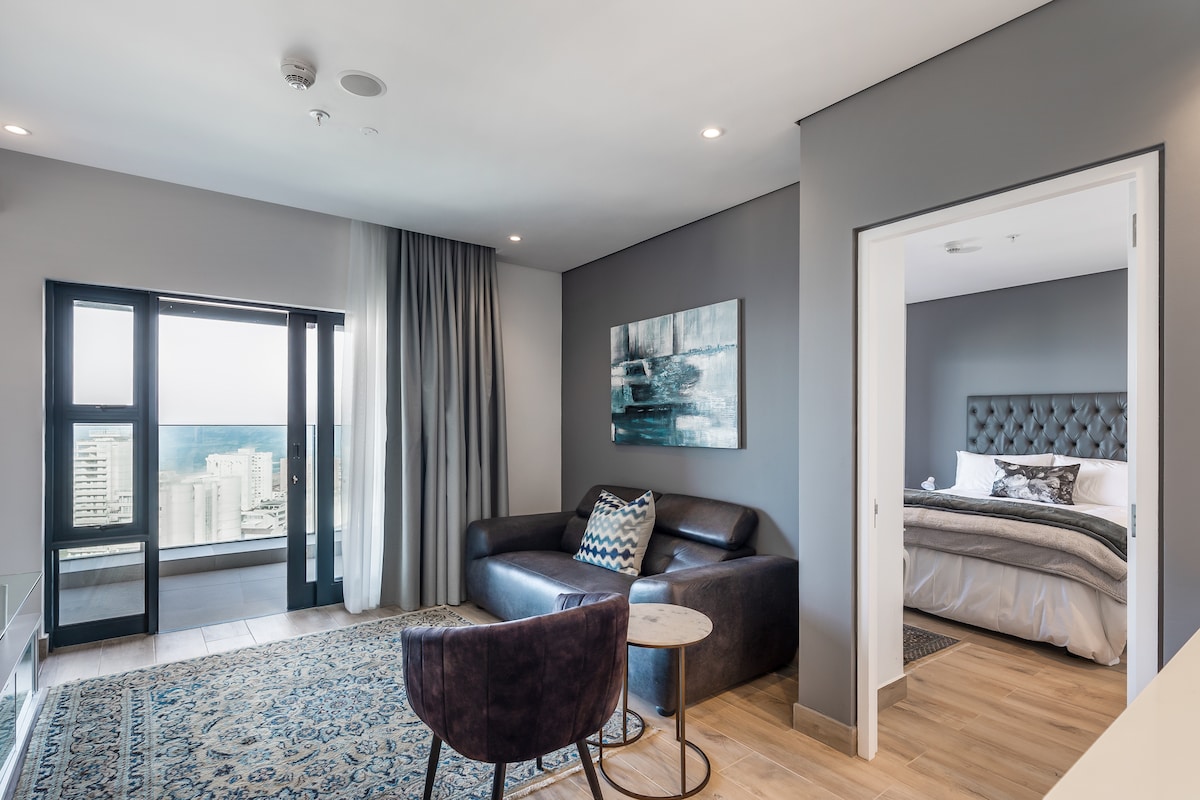 A contemporary living space is visible, featuring a dark sofa beside a small table. A large window offers a view of the surroundings. The bedroom is partially visible, showcasing a spacious bed with light bedding and headboard. Soft lighting enhances the neutral color scheme.