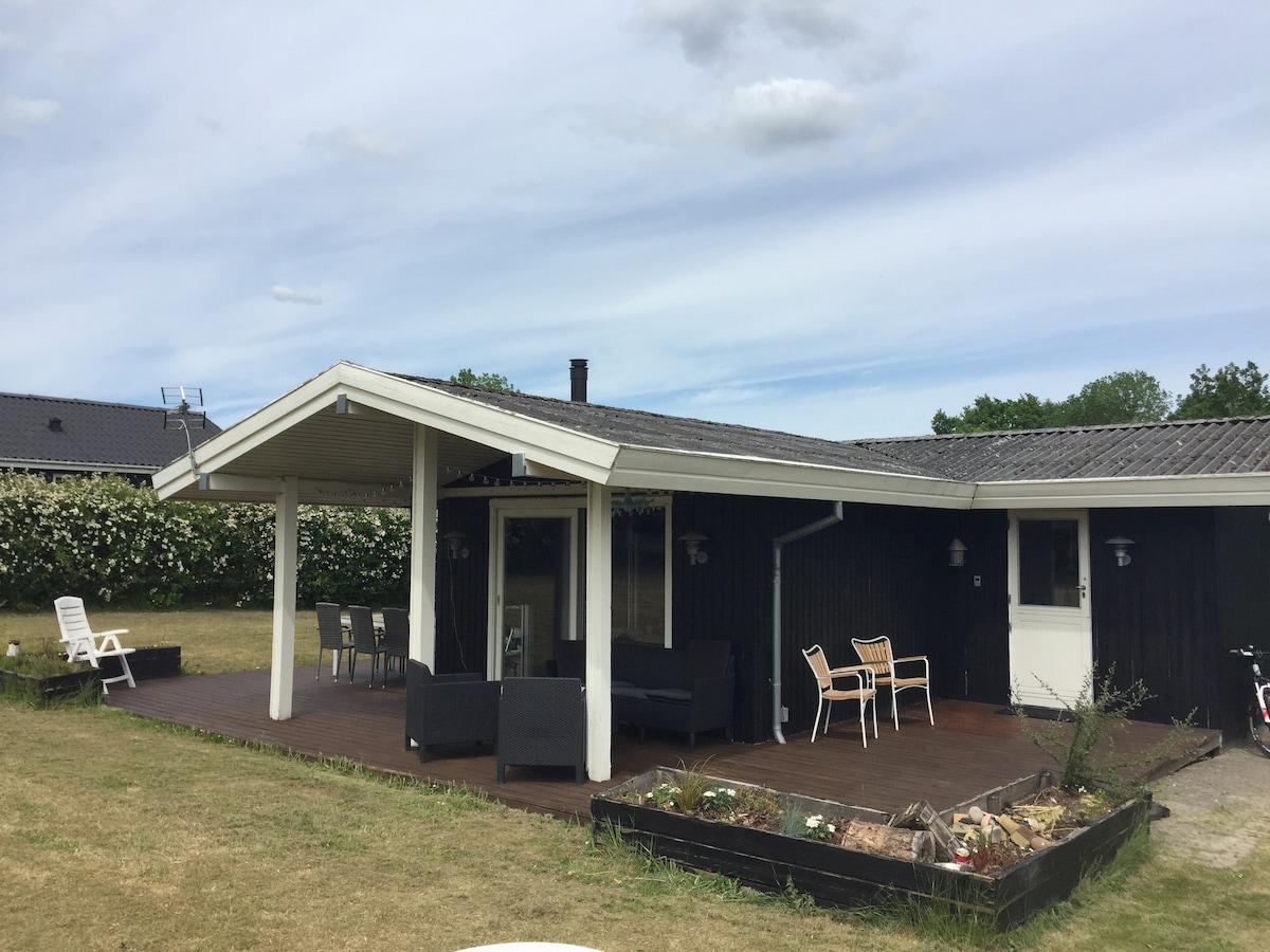 A wooden terrace extends from the house, furnished with several outdoor chairs and a table. A lawn surrounds the area, complemented by a flower bed showcasing various plants. The roof features an angled design, providing shelter over the seating area.