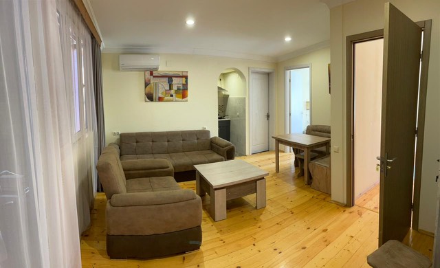 A spacious living area features a soft sectional sofa arranged around a wooden coffee table. Natural light filters through sheer curtains, illuminating the warm wood flooring. A dining table for four is positioned nearby, with a doorway leading to the kitchenette visible in the background.