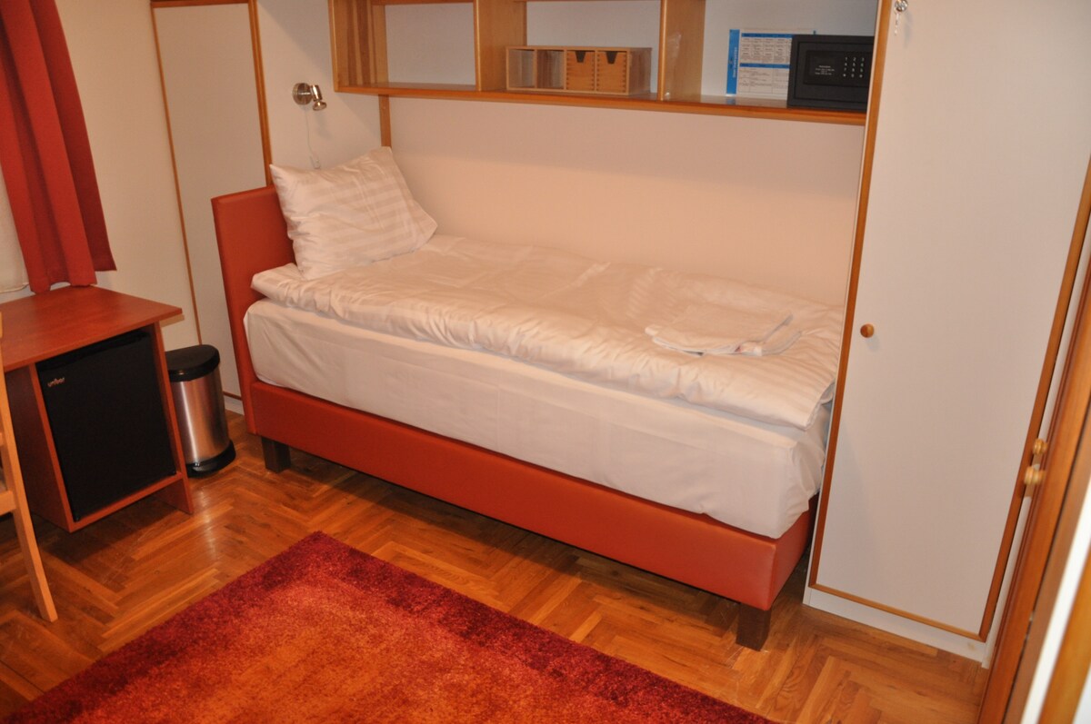 A neatly arranged sleeping area features a single bed with crisp white linens and a supportive headboard. A compact desk and a small waste bin are present, while a built-in shelf offers additional storage. The warm wooden floor is complemented by a deep red area rug.