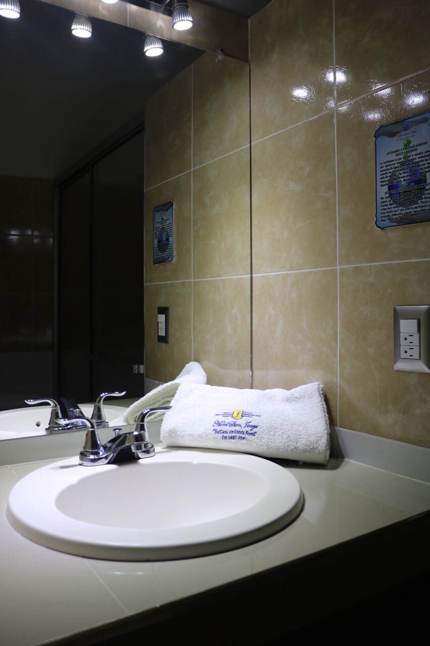 A well-lit bathroom features a round sink with modern fixtures and a neatly arranged white towel. The tiled walls reflect the soft glow from overhead lighting, adding to the clean ambiance of the space.