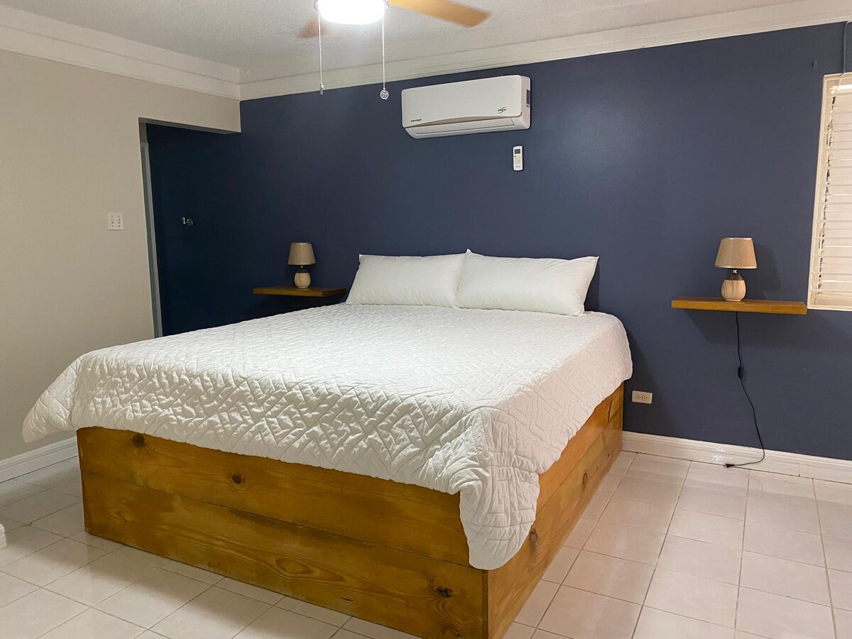 A spacious bed is centered against a deep blue accent wall, topped with crisp white linens. Nightstands with soft lamps are positioned on either side, while a ceiling fan provides ventilation. A window with blinds allows for natural light to fill the room.