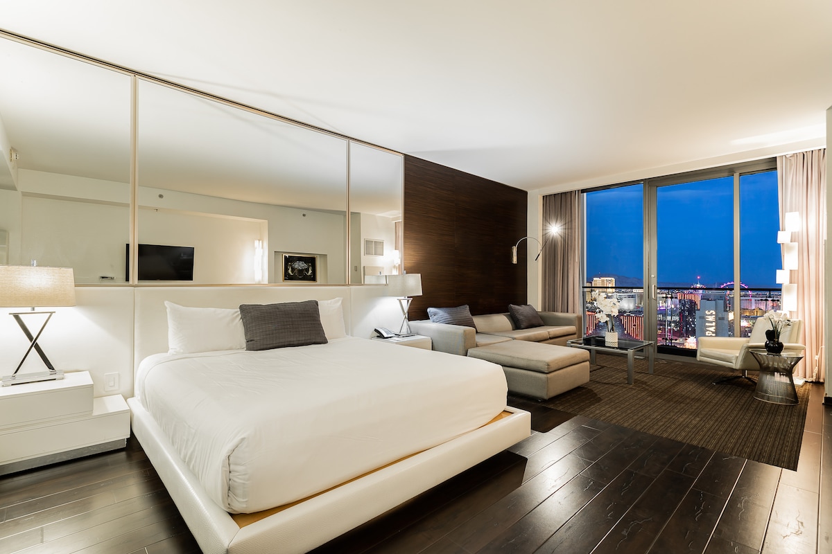 A spacious room features a large, inviting bed with crisp white linens, complemented by a modern sectional sofa. Floor-to-ceiling windows offer expansive views of the city skyline, while soft lighting creates a calming atmosphere. Contemporary furnishings include a bedside lamp and sleek accent table.