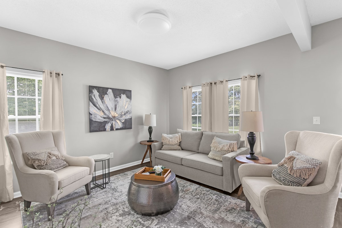 A bright living room features a light-colored sofa and two armchairs, creating a cozy seating arrangement. A coffee table with a metallic finish is centered on a patterned area rug. Natural light filters through the curtains of the windows, enhancing the serene atmosphere.