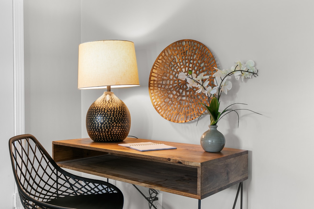A wooden desk is positioned against a light-colored wall, accompanied by a patterned lamp and a decorative round wall piece. A vase with an orchid and a notepad rests on the desk, alongside a stylish chair showcasing a geometric design.