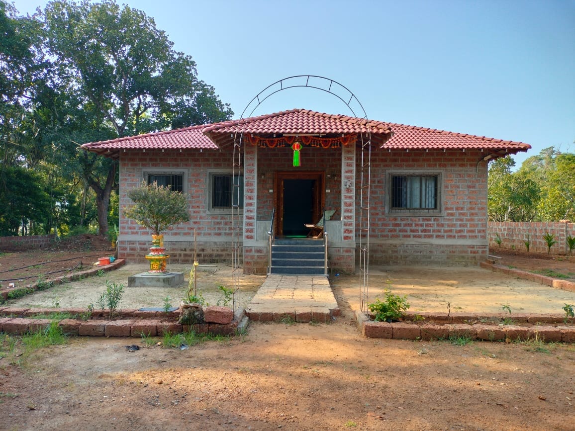 A single-story house is highlighted with a red-tiled roof and brick exterior. The entrance features a small set of steps leading to a doorway framed by decorative elements. Surrounding the home, a landscaped area includes patches of grass and small plants.