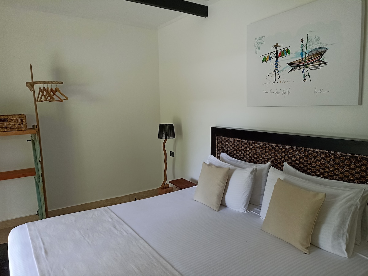 A spacious room features a queen size bed with crisp white linens and decorative pillows. A piece of wall art depicts a local fishing scene. A clothing rack and a lamp are positioned near the bed, adding functional elegance to the setting.