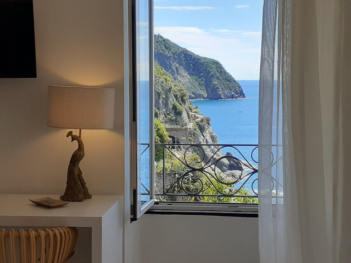 A window showcases a stunning sea view, framed by light, airy curtains. A lamp with a decorative base is positioned on a small table nearby, while the tranquil blue waters and surrounding greenery create a calming ambiance.