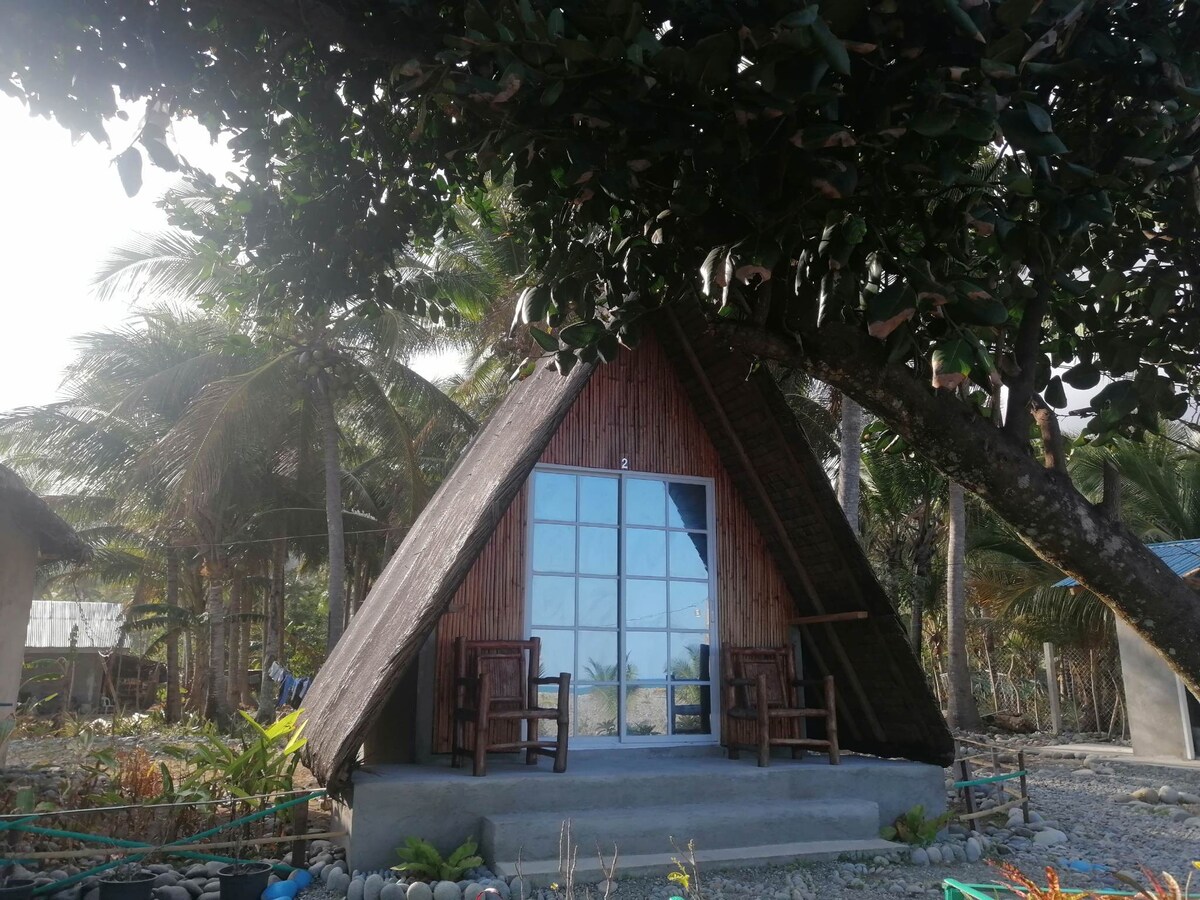 A charming teepee-style cottage is nestled among palm trees, featuring a large glass-paneled door. Two wooden chairs are placed on the outdoor porch, offering a cozy spot to enjoy the surroundings. The natural landscape creates a serene atmosphere by the beachfront.