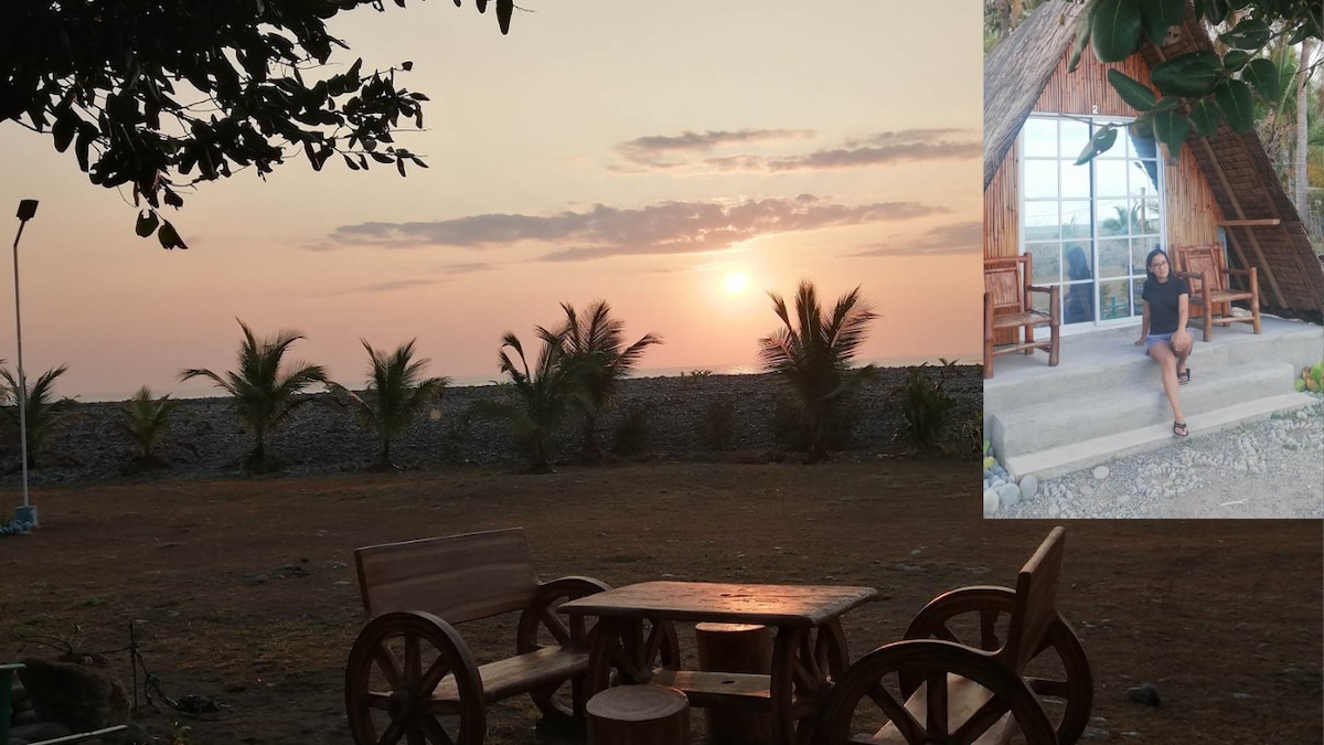 The image captures a serene sunset over the ocean, with the sun dipping below the horizon and palm trees outlined against the colorful sky. A rustic wooden table and chairs are positioned nearby, inviting relaxation in this tranquil outdoor space adjacent to the teepee.