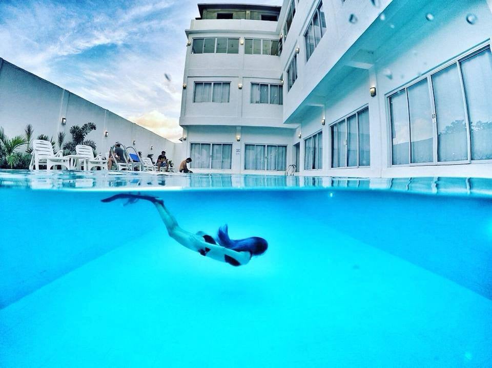 A clear swimming pool showcases a swimmer gliding beneath the surface. Sunlight reflects off the water, revealing the hotel building with large windows in the background. Lounge chairs are positioned nearby, creating a relaxed atmosphere for guests.