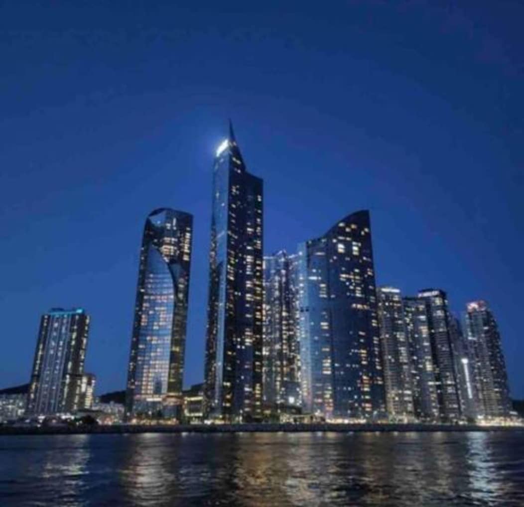 A modern skyline is illuminated against the night sky, showcasing tall buildings with reflective glass facades. The water in the foreground captures the city lights, creating a vibrant atmosphere. Various architectural designs are visible, highlighting the contemporary urban landscape.