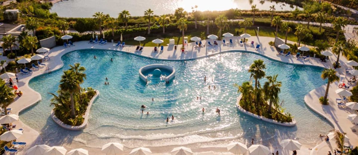 A sprawling pool area features gentle waves and a sandy beach-like feel, surrounded by palm trees. Sunshades and lounge chairs align along the edges. People can be seen enjoying the water, while the sun casts a warm glow over the tranquil setting.