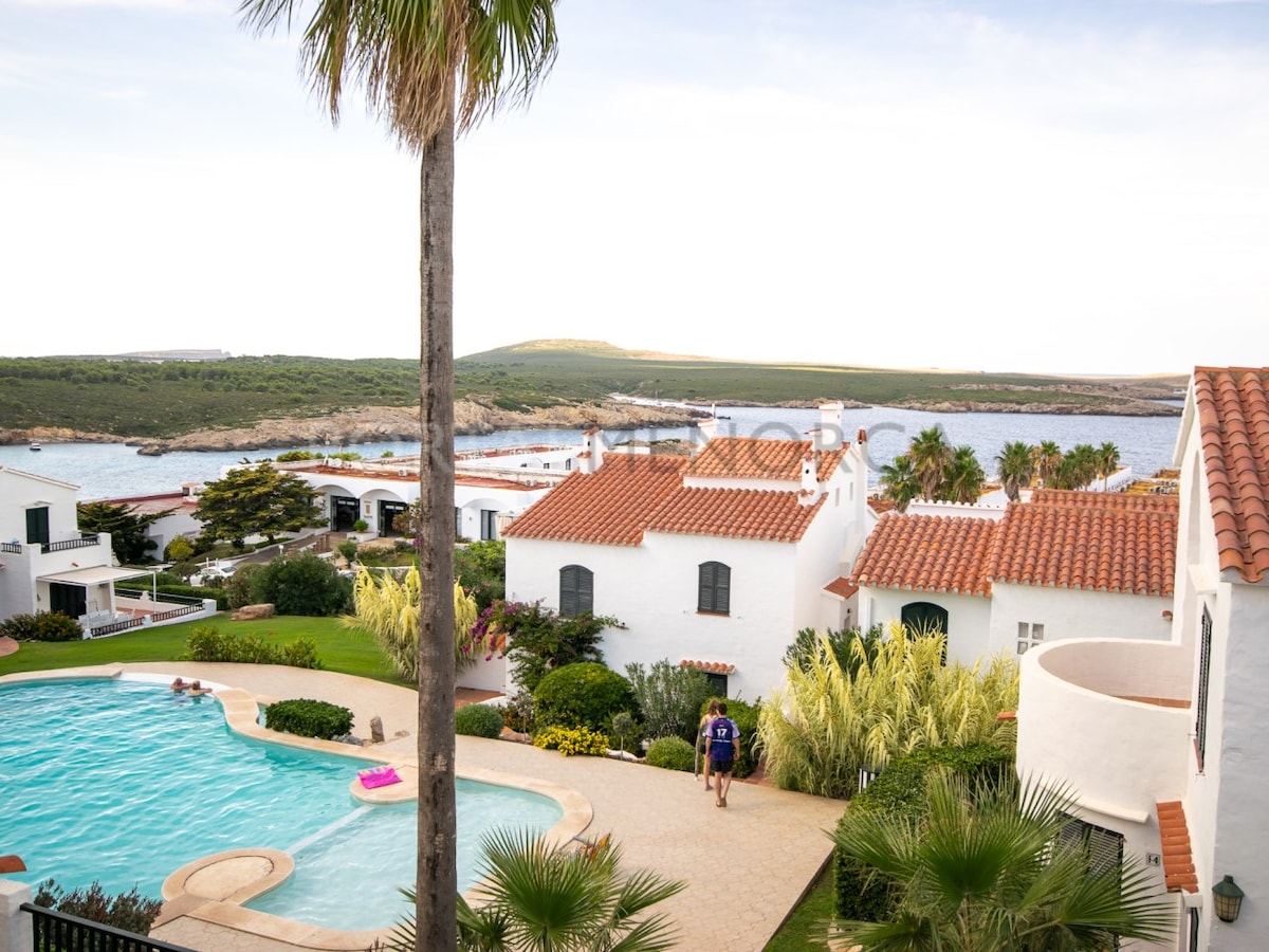 A view of the complex with whitewashed buildings featuring terracotta roofs is presented. A serene swimming pool takes center stage, surrounded by lush greenery. In the distance, the coastline and water are visible, complemented by the natural landscape.