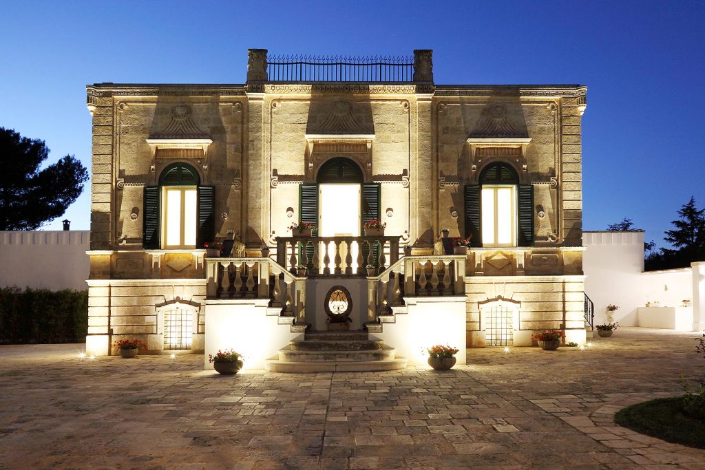 The historic villa is illuminated at night, showcasing its elegant facade with large windows. A grand staircase leads to the entrance, flanked by beautifully arranged planters. The architectural details reflect a classic style, contributing to the villa's sophisticated appearance.