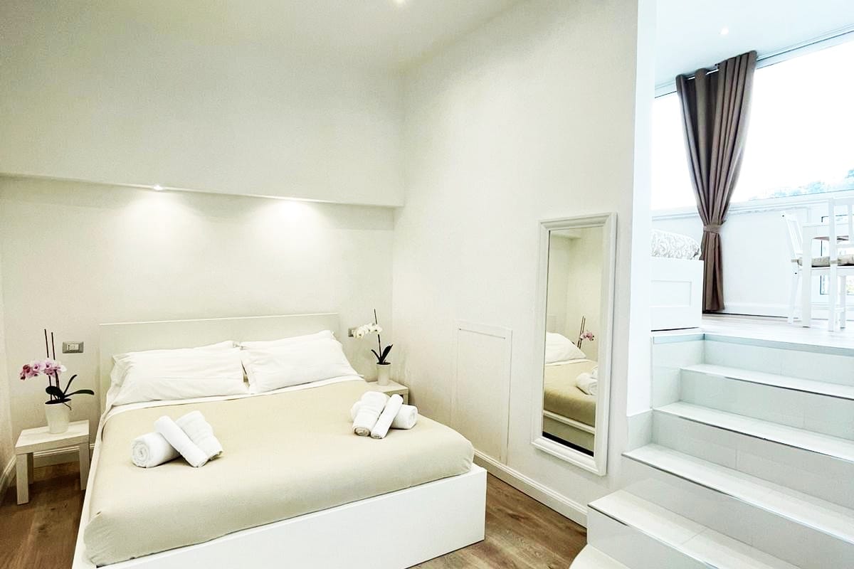A bright bedroom features a spacious bed with a neutral-colored blanket, flanked by twin side tables. Light is provided by recessed ceiling fixtures, and a full-length mirror reflects the inviting space. A staircase leads to an area with additional seating, complemented by natural light from a large window.