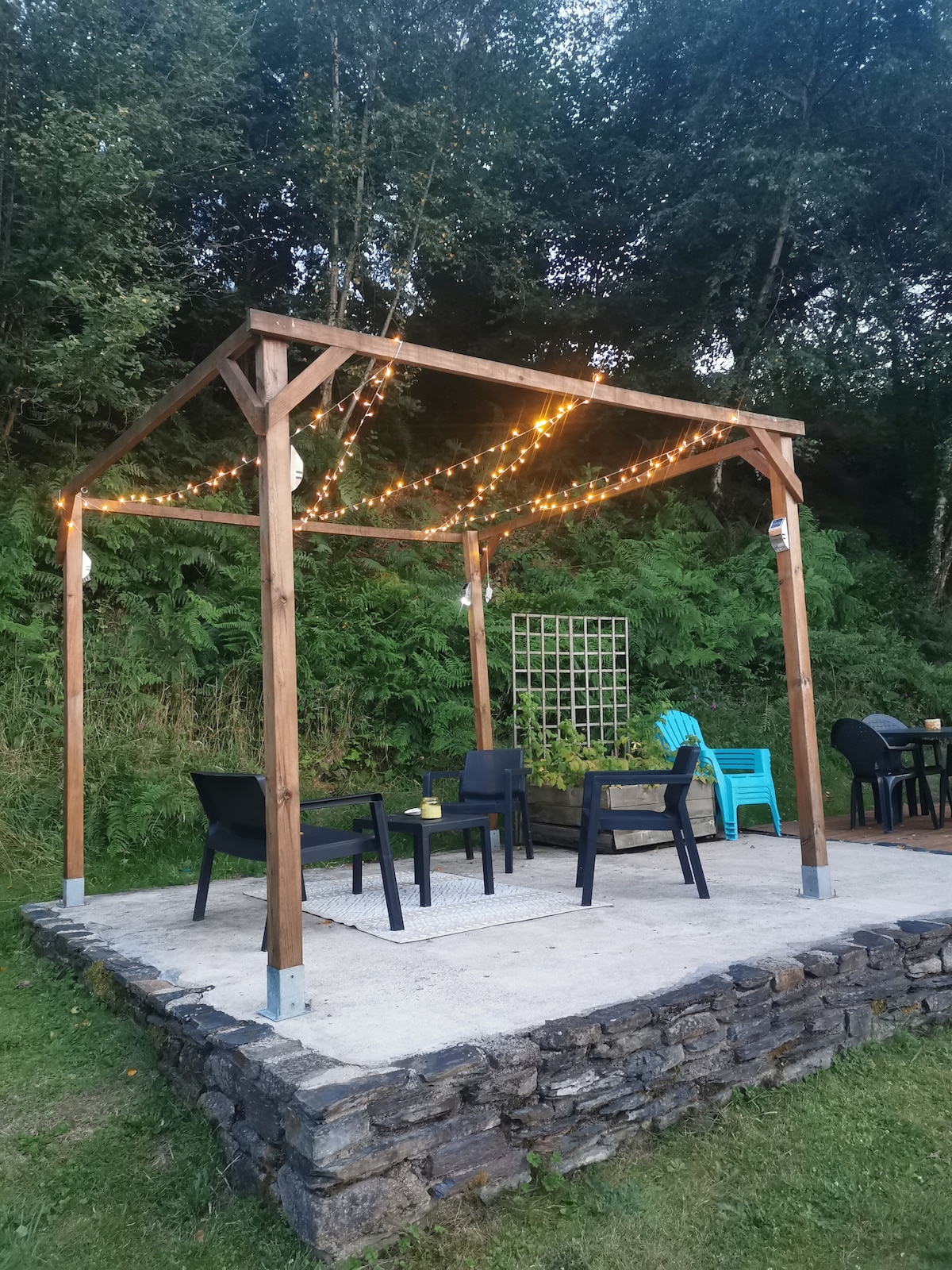 A wooden gazebo is adorned with string lights, creating a warm ambiance. Underneath, black chairs are arranged around a small table, alongside a bright blue chair. The setting is surrounded by greenery, enhancing the outdoor relaxation area.