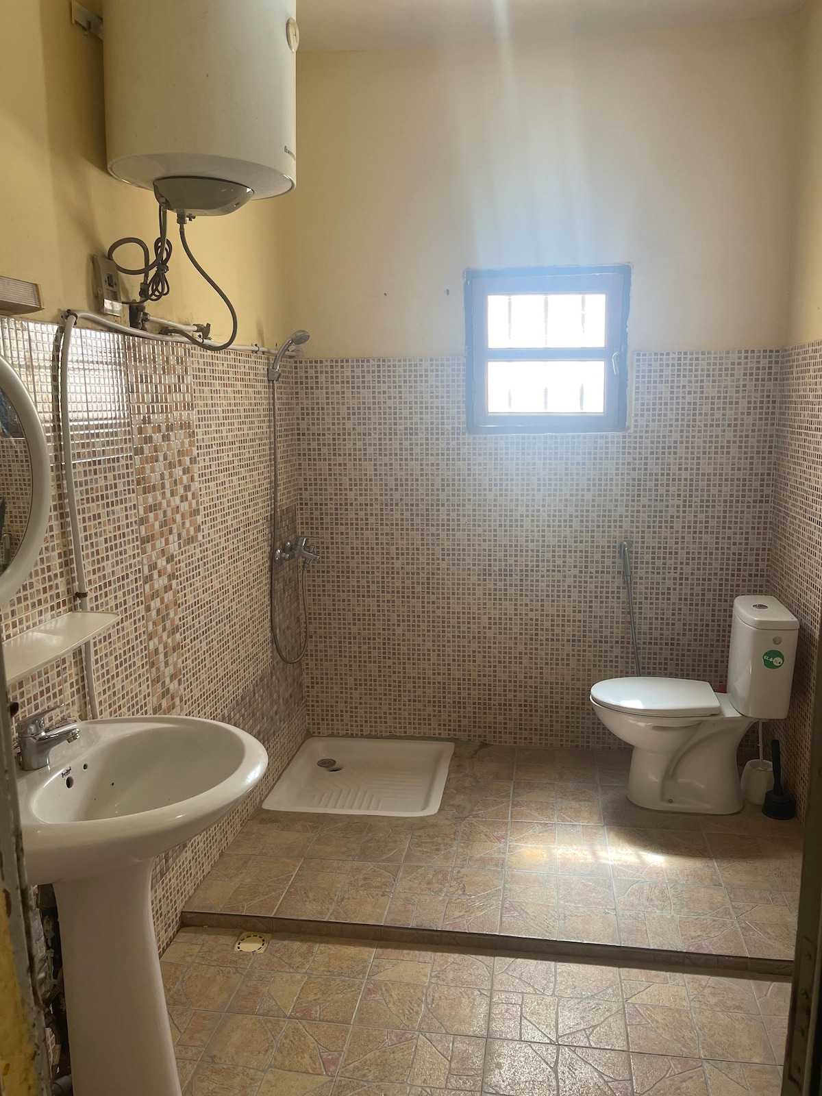 The bathroom features a shower area with tiled walls and a floor-mounted toilet. A window allows natural light to enter, and a sink is positioned beside a mirror. The overall layout highlights a functional and practical space.