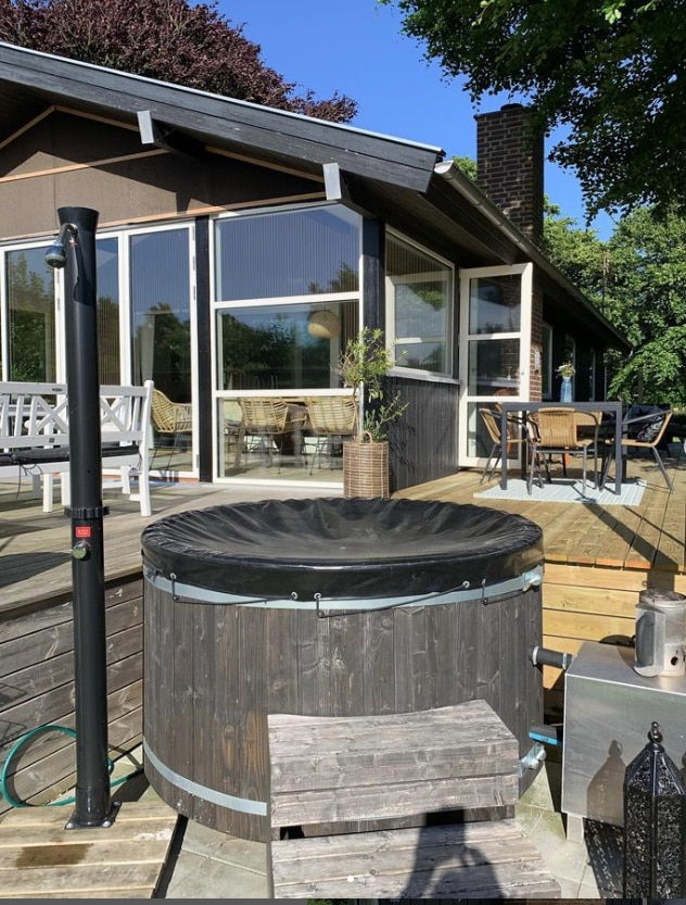 A wooden hot tub is positioned on a spacious deck, accompanied by a metal stove. The deck features a combination of lounge seating and a dining area, with large windows of the house providing a view of the outdoor space. Surrounding greenery adds a touch of nature.