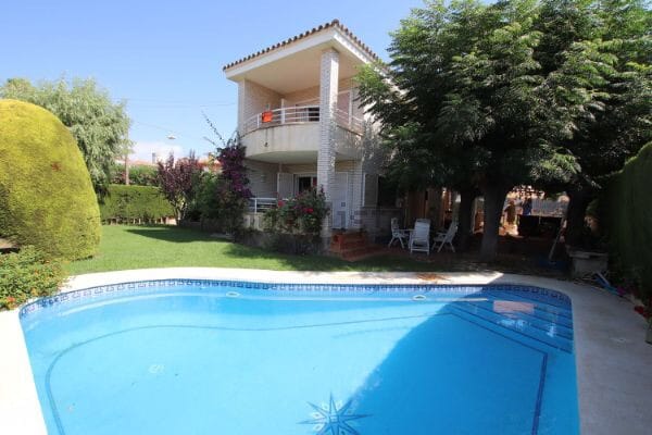 The villa is surrounded by lush greenery and features a private swimming pool with clear blue water. A spacious terrace is accessible from the upper level, and comfortable seating is available near the pool. The exterior is well-maintained, highlighting a relaxing outdoor environment.