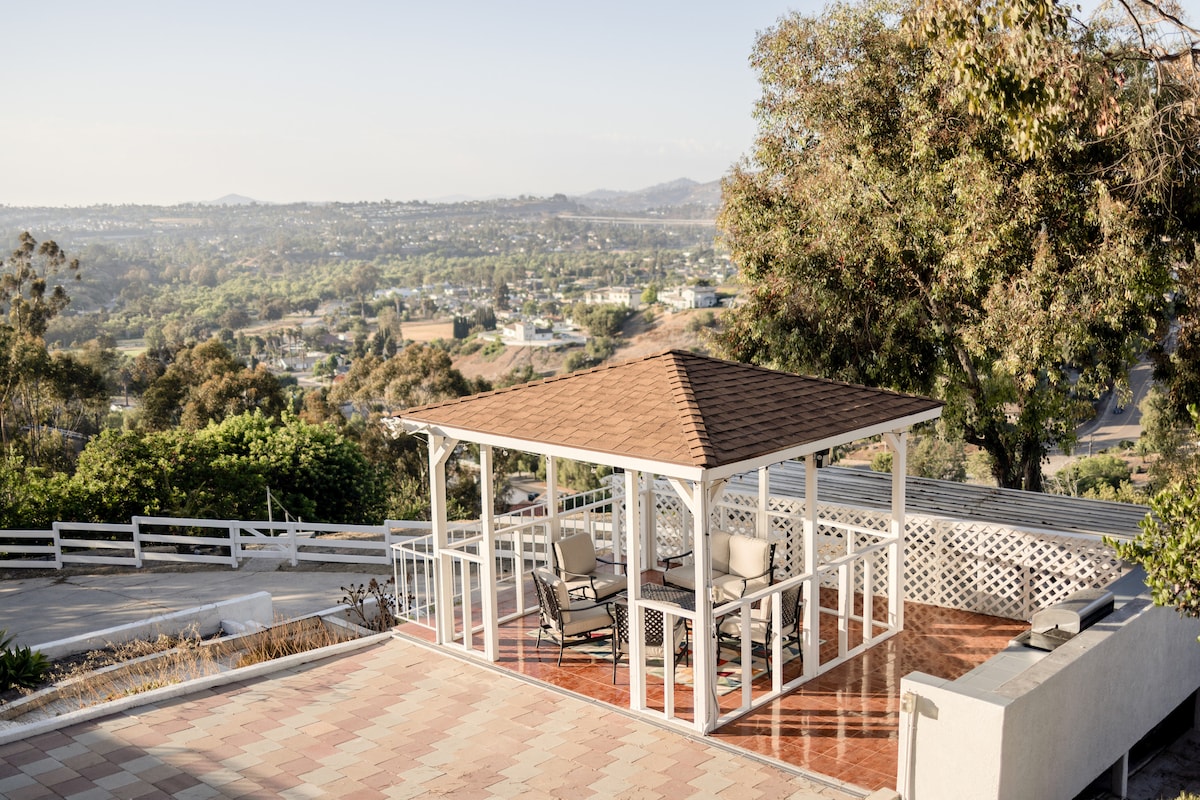A gazebo with a brown roof is set on a wooden deck, surrounded by lush greenery and expansive views of the hills and valleys. Comfortable seating is provided, creating an inviting space for relaxation and outdoor gatherings.