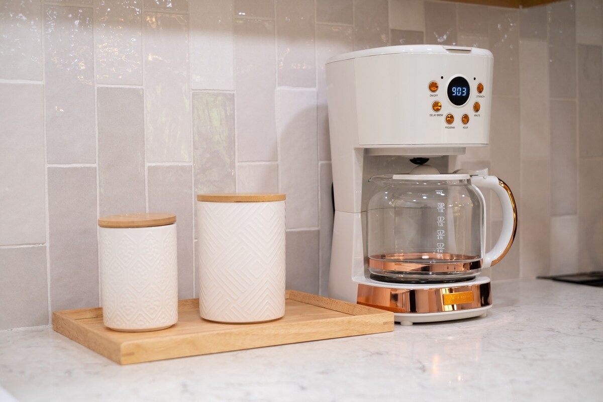A modern kitchen countertop is displayed with a coffee maker and two decorative storage containers on a wooden tray. The soft tones of the ceramic containers contrast with the sleek finish of the coffee maker, all set against stylish, textured backsplash tiles.