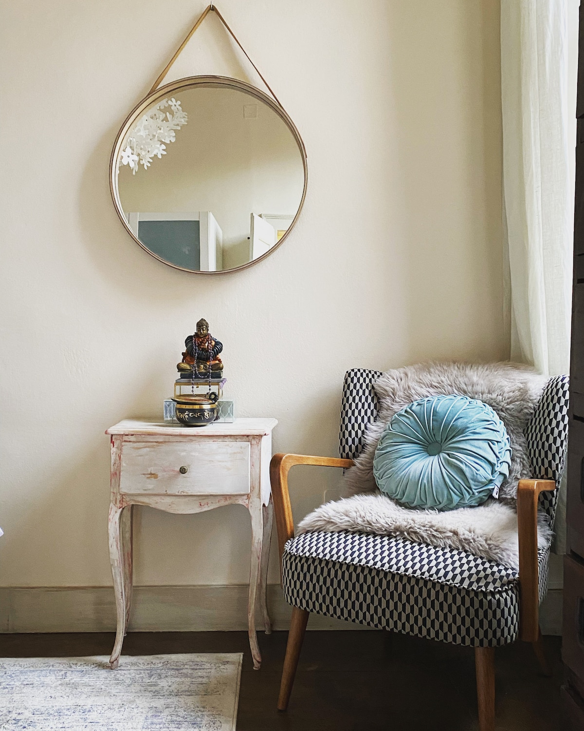 A seating area features a patterned armchair with a plush cushion and a cozy throw draped over the side. A small, distressed side table holds a stack of books and a decorative figure, with a round mirror reflecting light and enhancing the space.