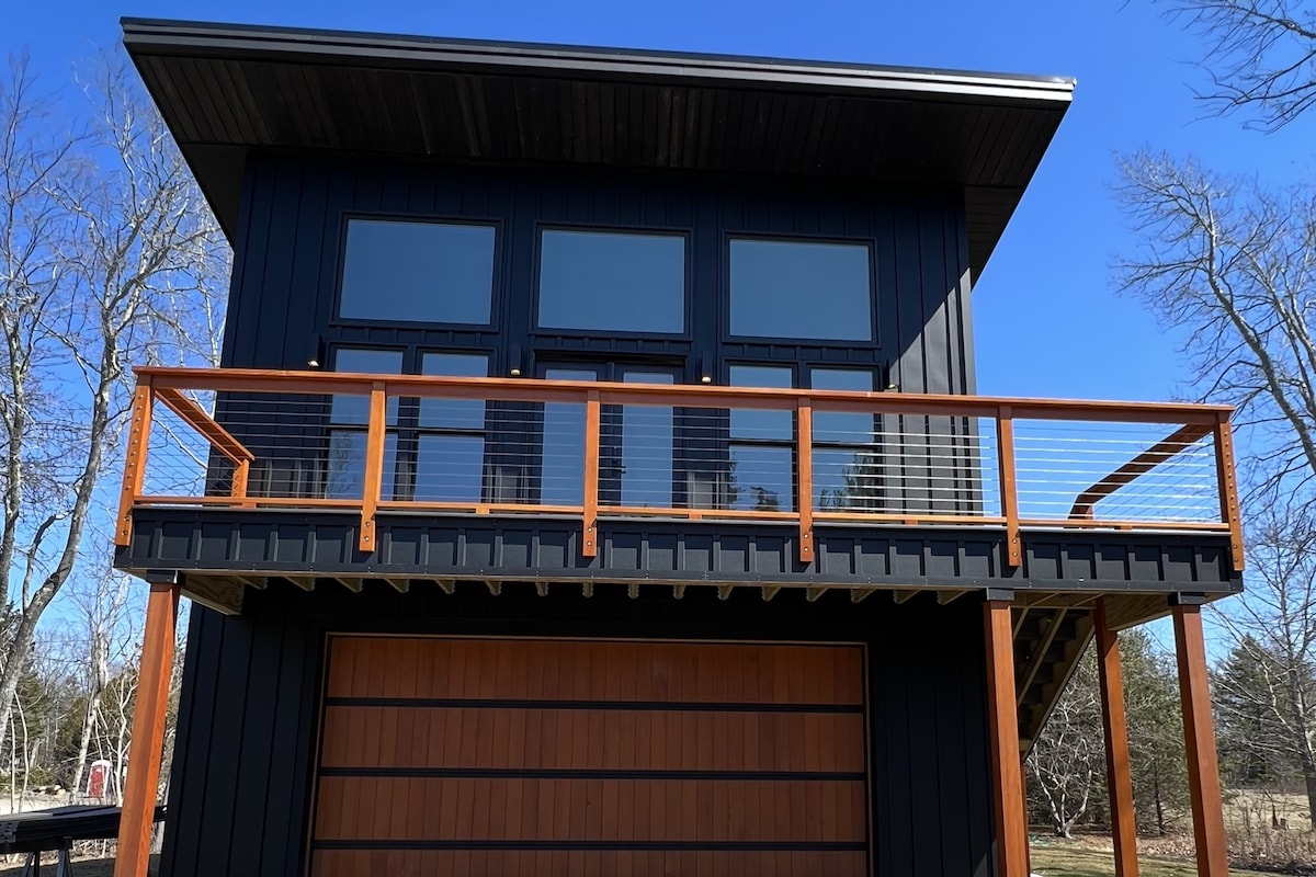The exterior of a modern two-story home is presented, featuring large windows across the upper level. A wooden deck with a railing extends from the front, while a garage door is situated below, showcasing a sleek design against a clear blue sky.