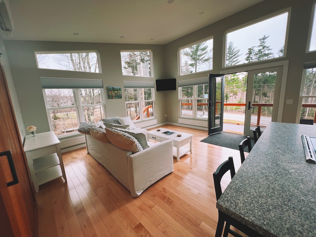 A bright living area features towering windows that overlook the surrounding trees. A light-colored sofa with patterned cushions is positioned opposite a coffee table. The hardwood floor reflects natural light, while a sleek kitchen island is visible in the foreground.