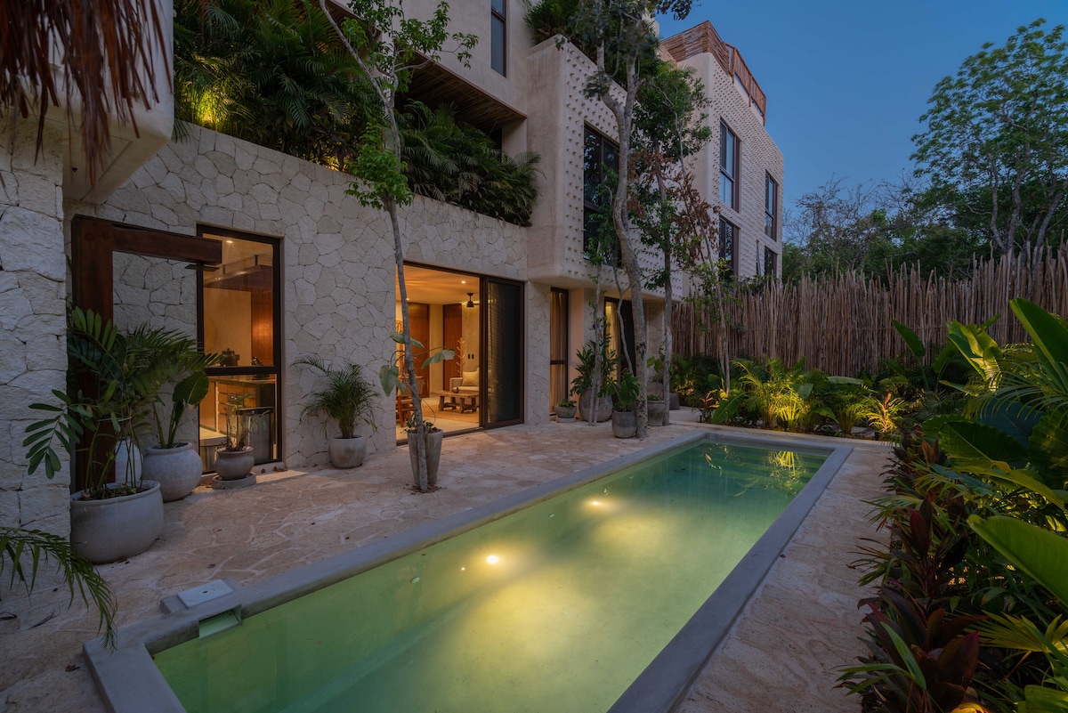 A serene garden space features a private pool, surrounded by lush tropical plants. Warm light illuminates the water, enhancing the tranquil atmosphere. Stone pathways lead to spacious glass doors, providing a seamless connection to the indoor living area.