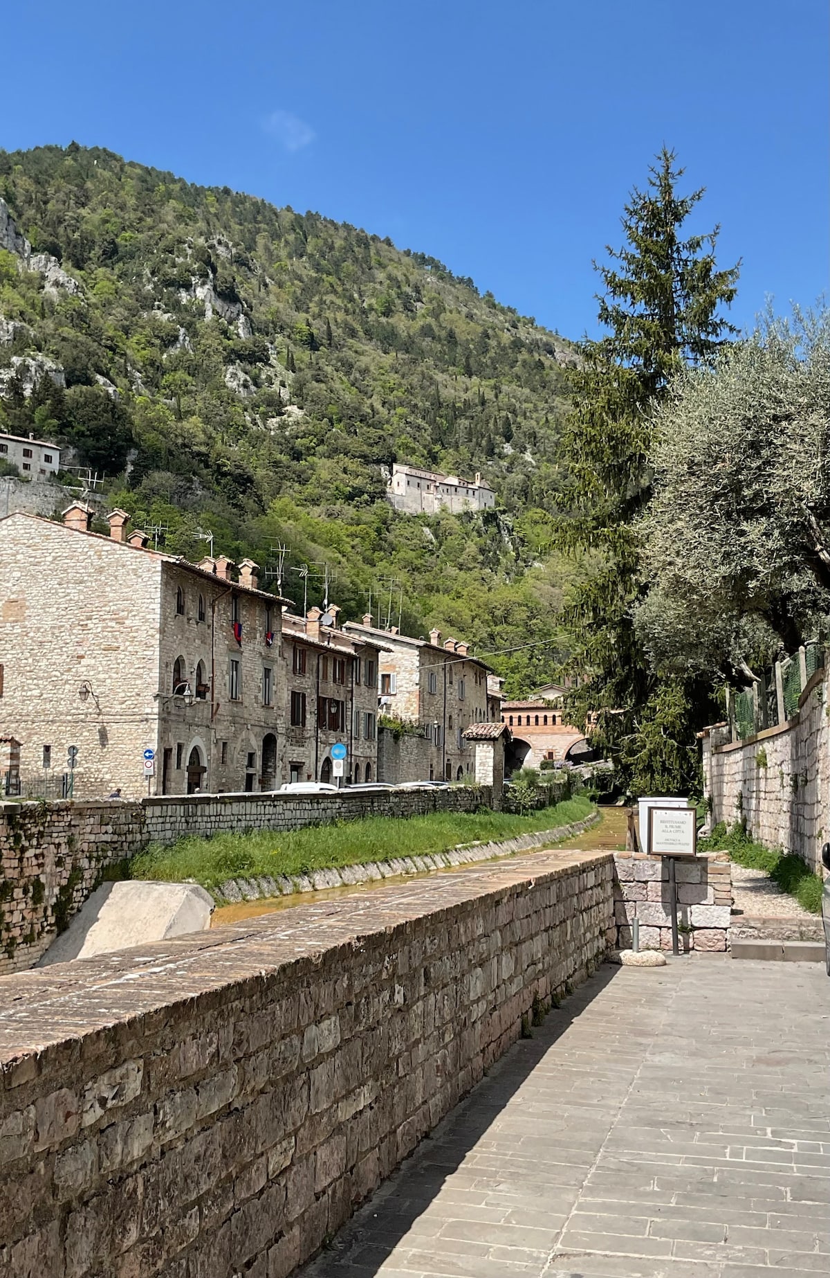 A serene street scene showcases historic stone buildings nestled against a lush green hillside. The landscape is complemented by trees and gentle slopes, while a pathway runs alongside a canal. The bright sky enhances the tranquil atmosphere of the setting.