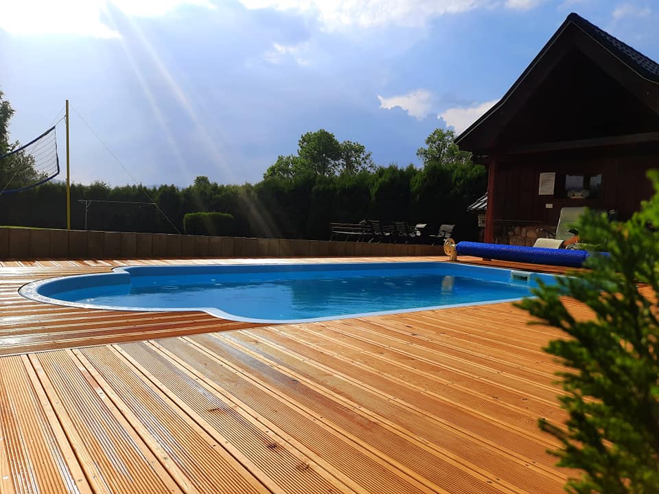 A clear blue pool is set on a polished wooden deck, surrounded by greenery. Sunlight reflects off the water's surface, while a volleyball net and outdoor seating area are visible in the background, enhancing the inviting atmosphere for relaxation.
