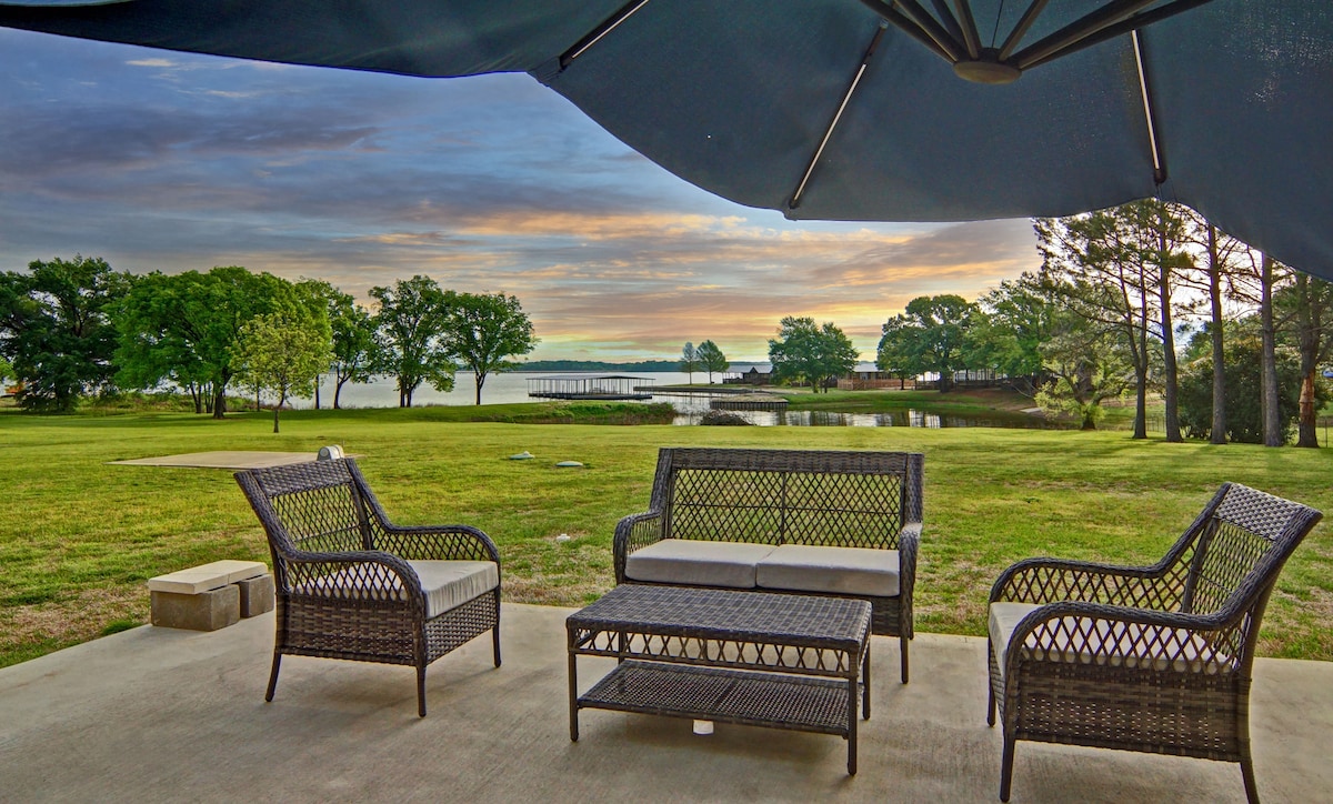 A shaded patio area features a set of four wicker chairs and a coffee table, arranged for relaxation. Lush green grass extends towards the serene waters of Lake Fork, with trees lining the shoreline and a glimpse of a dock visible in the distance beneath a colorful sky.