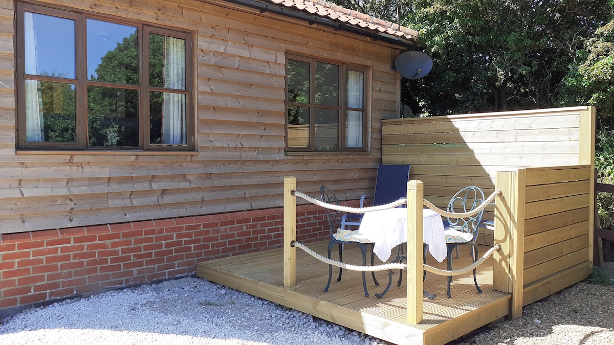A wooden deck area is shown, featuring a round table and four chairs, surrounded by a protective railing. The structure's exterior is clad in wooden panels, and two windows allow natural light to enter. A private space is created, providing an intimate setting.