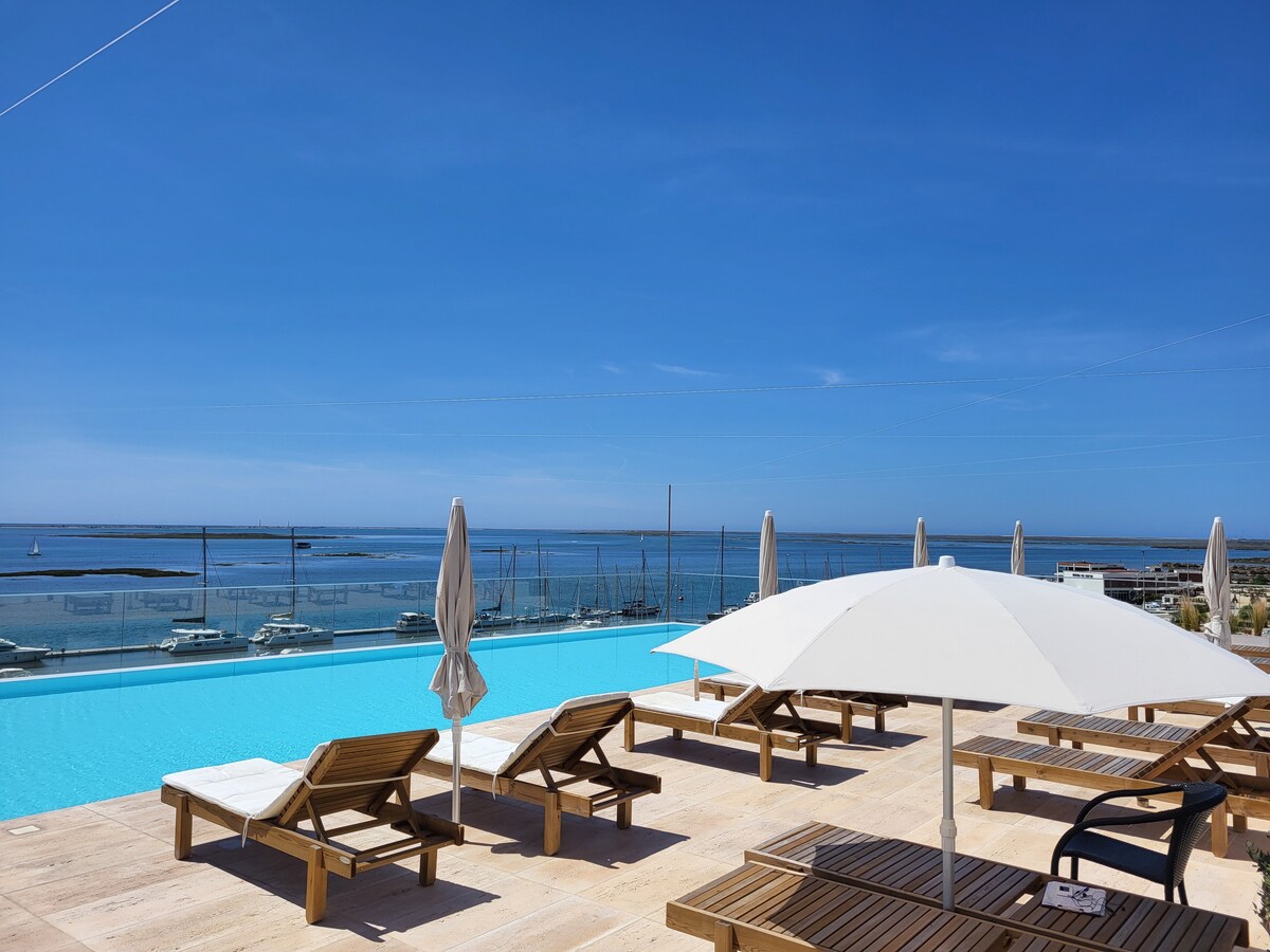An outdoor rooftop pool features a serene infinity edge that blends with the ocean view. Sun loungers line the pool area, accompanied by large umbrellas providing shade. The sky is clear with bright blue tones, enhancing the calm atmosphere of the scenery.