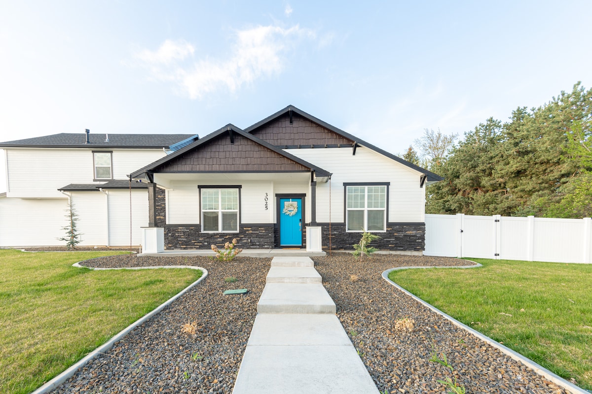 A modern home is showcased with a two-tone exterior combining dark shingles and light siding. The vibrant blue front door is framed by large windows. The lawn is neatly manicured, with a pathway of concrete leading to the entrance, bordered by decorative stones and small plants.