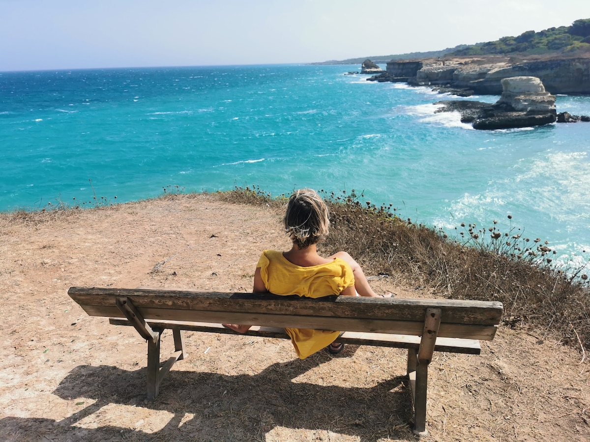 A wooden bench overlooks the coastline, with turquoise waters gently lapping against rugged cliffs. The scene captures a figure sitting on the bench, facing the expansive sea. Wild grasses are visible in the foreground, blending into the natural landscape.
