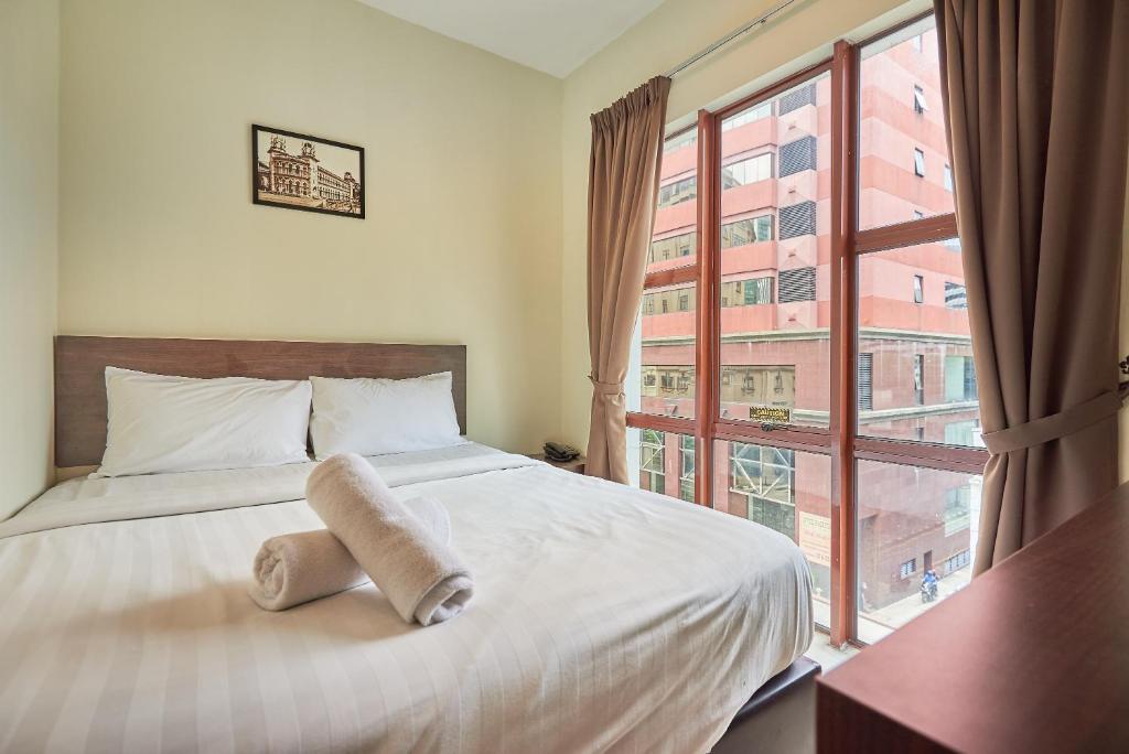 A queen bedroom is shown with a spacious bed covered in light linens, flanked by soft, neutral-colored curtains. A rolled towel is placed on the bed. Large windows offer a view of adjacent buildings, allowing natural light to fill the room.