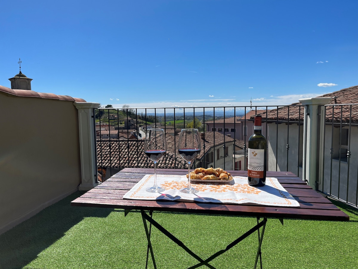A terrace table is set with two glasses of red wine and a bottle, complemented by a plate of pastries. The background showcases a clear blue sky and rolling hills, providing a serene outdoor space ideal for relaxation.
