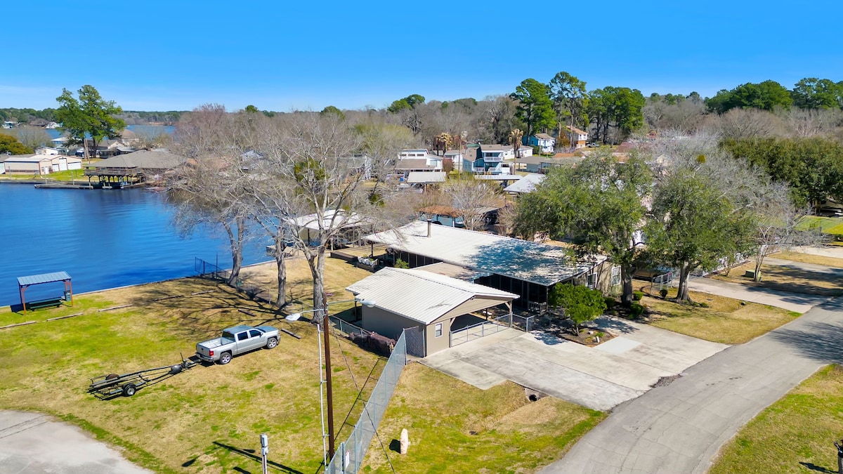 An aerial view showcases the exterior of Lee Shore Lake House surrounded by grassy areas and trees. The boat dock is visible along the lake's edge, with parked vehicles and a paved driveway leading to the entrance of the home.