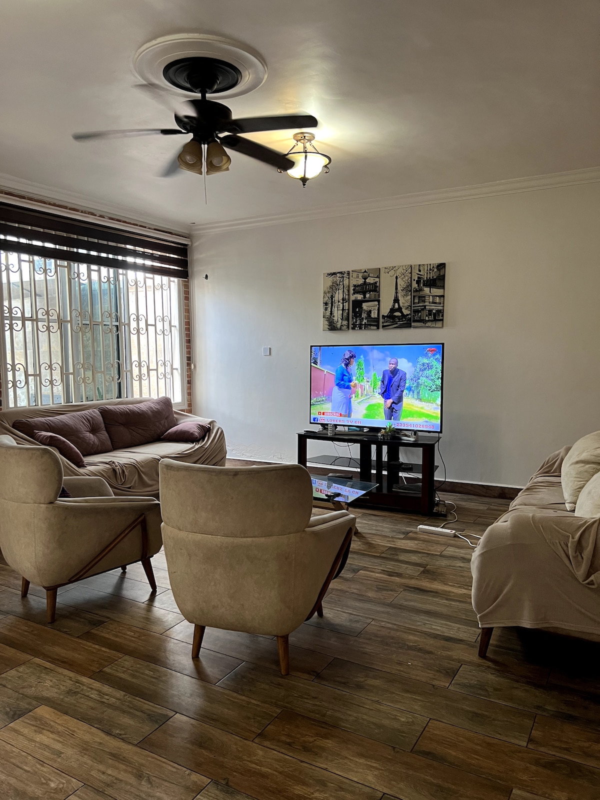 A cozy living area is presented with soft seating arrangements, including a pair of beige armchairs and a light-colored couch. A television is positioned on a low TV stand, displaying a program. Natural light enters through large windows with decorative grilles.